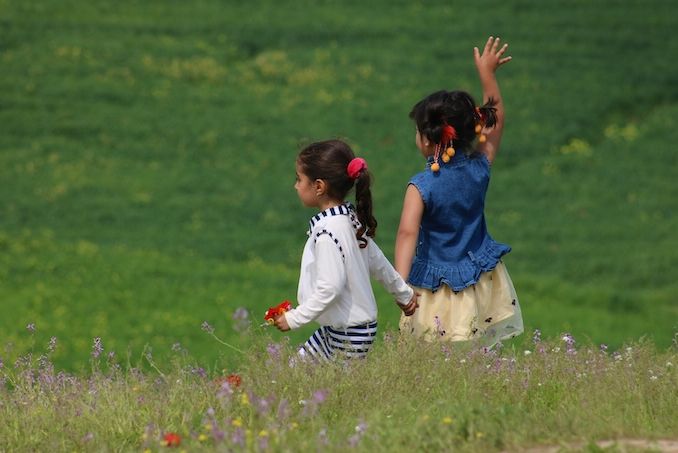 Photo Credit: Firoz Sidiqy. Young sisters holding hands in a green field facing away with one girl waving to someone off frame.