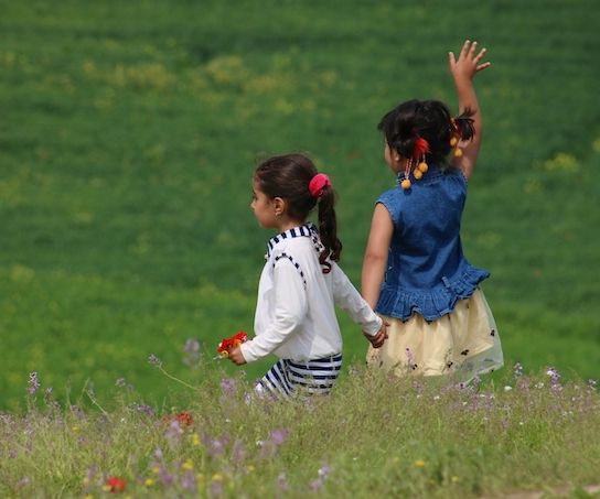 Photo Credit: Firoz Sidiqy. Young sisters holding hands in a green field facing away with one girl waving to someone off frame.
