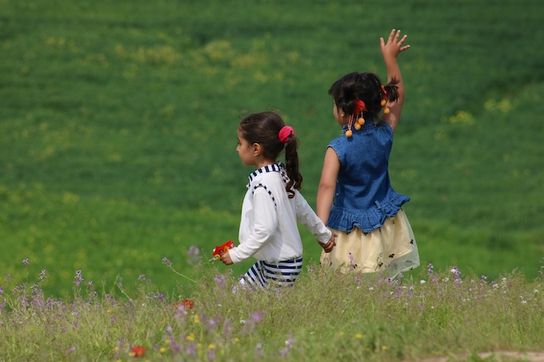 Photo Credit: Firoz Sidiqy. Young sisters holding hands in a green field facing away with one girl waving to someone off frame.
